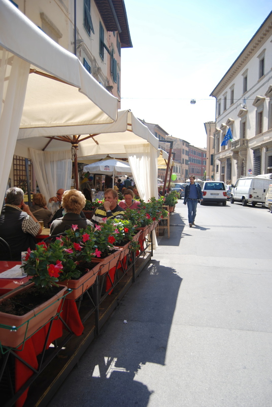 9 - A sunny street scene in an Italian town, featuring a sidewalk cafe with white canopy tents and planters of red flowers, alongside historic buildings and a person walking down the asphalt